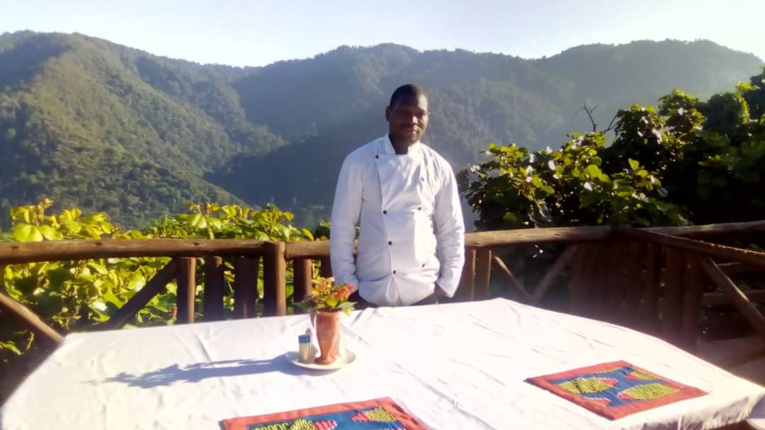 Executive Chef Kanaburenge Herbert at Orugano Bwindi Lodge, preparing locally sourced gourmet meals at an outdoor cooking station overlooking the lush Bwindi Impenetrable Forest—a highlight of the luxury Uganda safari dining experience